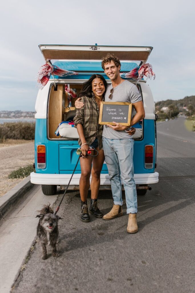 A smiling couple posing with their dog beside a classic van on a scenic road trip.